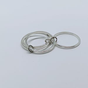 Puede incluir: Un conjunto de anillos plateados, que incluye tres anillos entrelazados y un anillo separado. Los anillos est&aacute;n hechos de un material met&aacute;lico brillante y est&aacute;n dispuestos sobre una superficie blanca. Probablemente sean joyas.