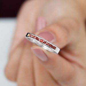 May include: A silver ring with a row of small, square, red gemstones. The ring is held up close, showing the details of the gemstones and the metal band. The gemstones are evenly spaced and set into the band.