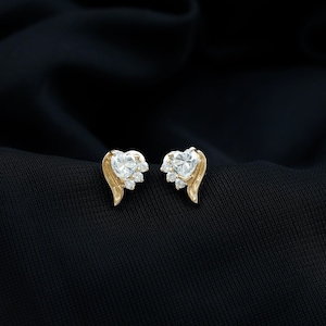 May include: A pair of gold-toned heart-shaped stud earrings. Each earring features a large, clear, heart-cut stone at the center, surrounded by smaller, clear stones. The earrings have a gold-colored wing-like design on the sides.