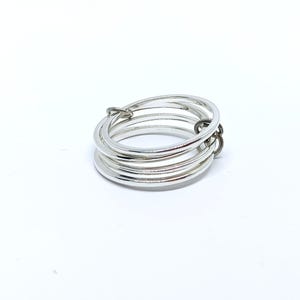 Puede incluir: Un anillo plateado con m&uacute;ltiples bandas, conectado por un peque&ntilde;o cierre de color oscuro. El anillo se presenta sobre un fondo blanco liso, resaltando su brillo met&aacute;lico y dise&ntilde;o minimalista. Las bandas del anillo son lisas y reflectantes.