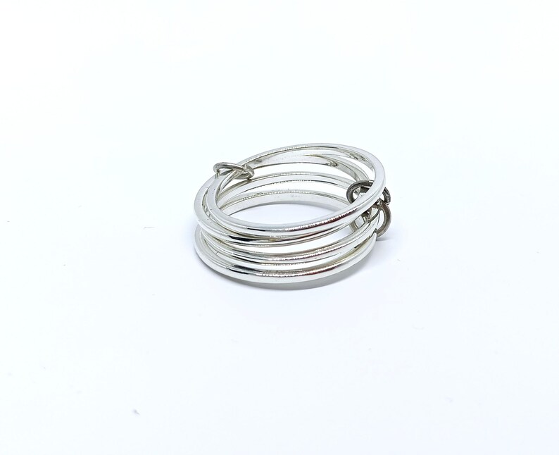 Puede incluir: Un anillo plateado con m&uacute;ltiples bandas, conectado por un peque&ntilde;o cierre de color oscuro. El anillo se presenta sobre un fondo blanco liso, resaltando su brillo met&aacute;lico y dise&ntilde;o minimalista. Las bandas del anillo son lisas y reflectantes.