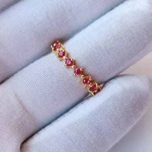 May include: A gold-colored ring with a series of small, round, red gemstones set in a row. The ring is displayed against a white background, highlighting the vibrant red of the gemstones and the ring's design.