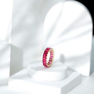 May include: A gold ring with a continuous band of rectangular, vibrant pink gemstones. The ring is displayed on a white pedestal against a minimalist white backdrop, highlighting the jewelry's design and color.