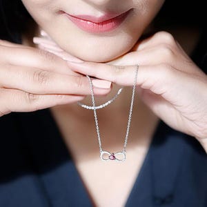 May include: A silver necklace featuring an infinity symbol pendant. The pendant is encrusted with small, clear stones and a central, heart-shaped, pink gemstone. The necklace is displayed against a dark blue background.