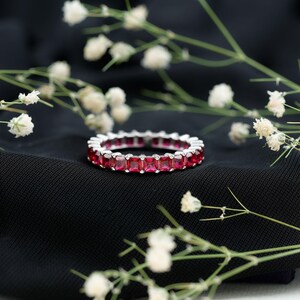 May include: A silver ring with a continuous row of square, deep red gemstones. The ring is set against a black background, with delicate white flowers in the foreground. The gemstones are evenly spaced around the band.