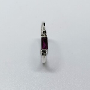 May include: A silver ring featuring a rectangular, deep red gemstone. The ring has a minimalist design with two small, round accents on either side of the gemstone. The band is thin and polished, creating a sleek and elegant look.
