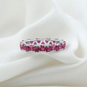 May include: A silver ring with a continuous band of heart-shaped, vibrant pink gemstones. The ring is set against a soft, white fabric background, highlighting the sparkle and color of the gemstones. A piece of jewelry.