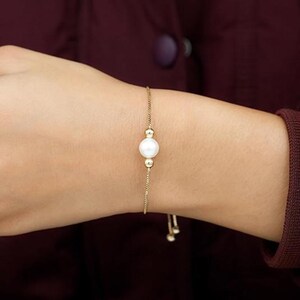 May include: A delicate gold bracelet featuring a large, round, white pearl as its centerpiece. The bracelet has a thin, adjustable chain with small gold beads. The pearl is the focal point, creating a simple yet elegant design.