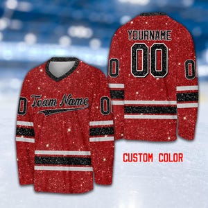 May include: Red and black hockey jersey with a glitter finish. The jersey has a white stripe with black trim around the neck and sleeves. The jersey has the text "Team Name" and a number "00" with the text "YOURNAME" above it.