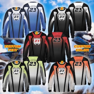 Puede incluir: Camisetas deportivas de manga larga en varias combinaciones de colores, incluyendo azul, negro, rojo, verde y naranja. Cada camiseta presenta un gráfico de escudo con el número 23 y espacio para texto personalizado. Las camisetas están diseñadas para deportes.