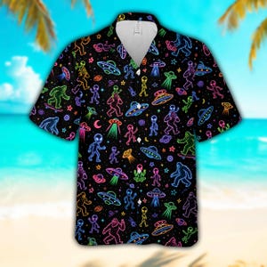 May include: A short-sleeved button-down shirt in black featuring a vibrant, neon-colored all-over print of Bigfoot figures, extraterrestrial beings, flying saucers, and stars. The shirt has a classic collar and a white inner neck lining.