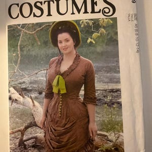 May include: A McCall's Costumes sewing pattern envelope M8189, size A5 (6-8-10-12-14). The cover features a brown dress with a ruffled neckline, three-quarter sleeves, a green bow at the chest, and a gathered skirt, worn with a green bonnet.