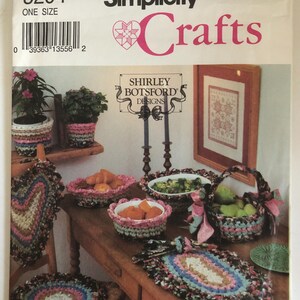 May include: A vintage Simplicity Crafts pattern cover featuring a variety of crocheted items. The image showcases a table with crocheted baskets, placemats, and a chair seat cover. The cover includes the text "Simplicity Crafts" and "Shirley Botsford Designs."