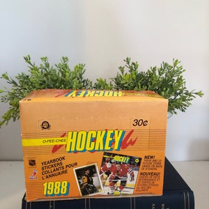 May include: A box of 1988 O-Pee-Chee Hockey trading cards. The box is orange and yellow with the words "O-Pee-Chee Hockey" in large, bold letters. The box also features a picture of a hockey player in a red and white jersey. The text "Yearbook Stickers Collants Pour L'Annuaire 1988" is printed on the box. The box is priced at 30 cents.