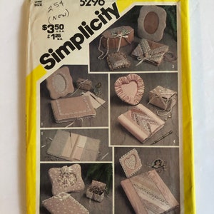 May include: Simplicity sewing pattern number 5296 for making various decorative boxes and a photo album. The pattern includes instructions for making a heart-shaped box, a square box, a round box, and a photo album. The pattern is on a yellow background with a white border.