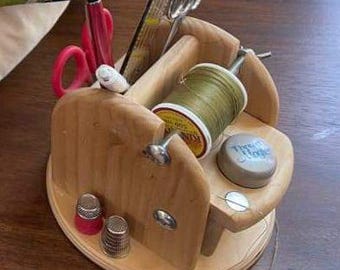 Puede incluir: Un organizador de costura de madera con un carrete de hilo, dos dedal, unas tijeras, un bolígrafo y una regla. El organizador tiene un pequeño compartimento para guardar los materiales de costura.