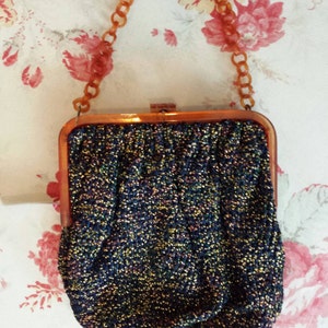 May include: Vintage evening bag with a shimmering, textured fabric in shades of blue, gold, and purple. The bag has a rectangular frame and a chain handle made of amber-colored plastic links. The clasp is a simple metal closure.