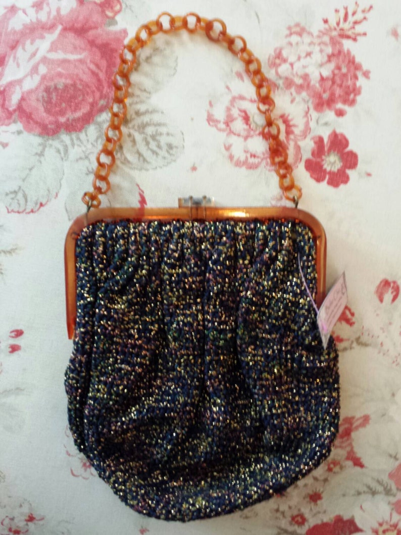 May include: Vintage evening bag with a shimmering, dark blue fabric, accented with gold and other colors. The bag has a tortoiseshell-style frame and handle. The clasp is silver-toned. The background is a floral pattern.