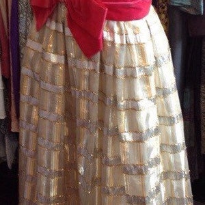 K&ouml;nnte beinhalten: Ein wei&szlig;es und goldenes Paillettenkleid mit einem rosafarbenen Satinband. Das Kleid hat ein tailliertes Oberteil und einen weiten Rock.