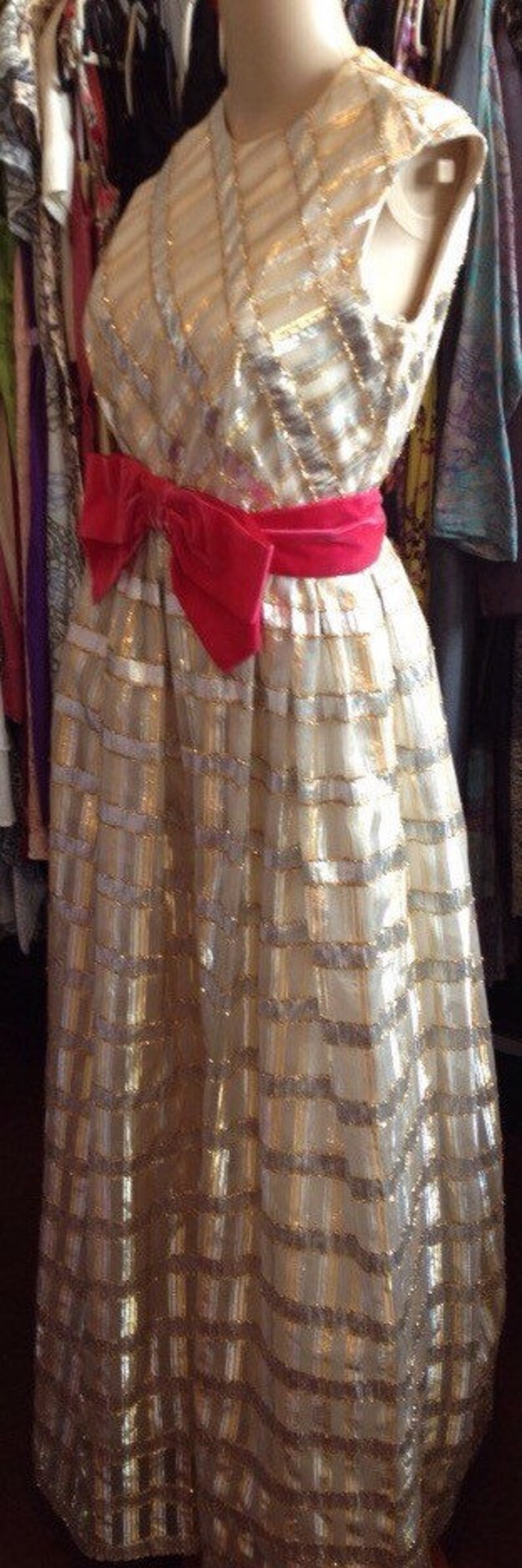 K&ouml;nnte beinhalten: Ein wei&szlig;es und goldenes Paillettenkleid mit einem rosafarbenen Satinband. Das Kleid hat ein tailliertes Oberteil und einen weiten Rock.