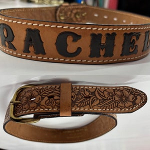 May include: A brown leather belt with the name "RACHEL" embossed in black letters. The belt has a floral design and a brass buckle.