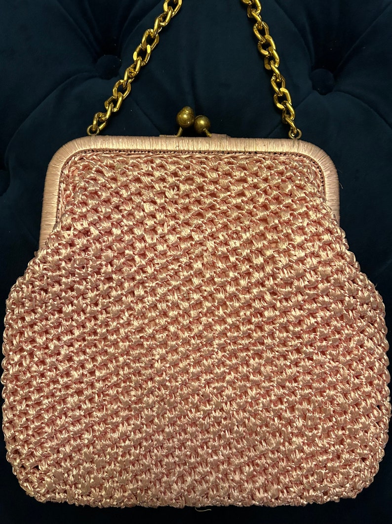 May include: A pink crocheted clutch bag with a gold chain strap and a brass clasp.