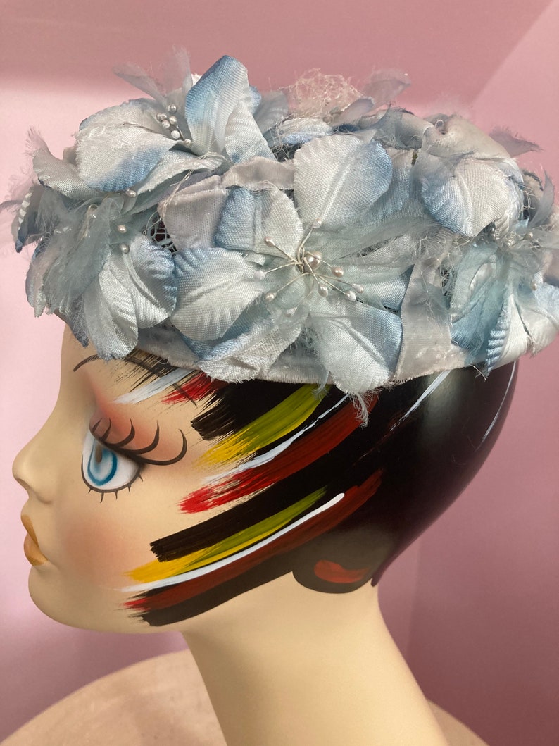 May include: A light blue hat with a flower design. The hat is made of fabric and has a net-like material on top. The hat is sitting on a mannequin's head.