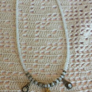 May include: A white beaded necklace with a gold cherub pendant and two smaller gold accents with white stones. The necklace is on a crocheted surface.