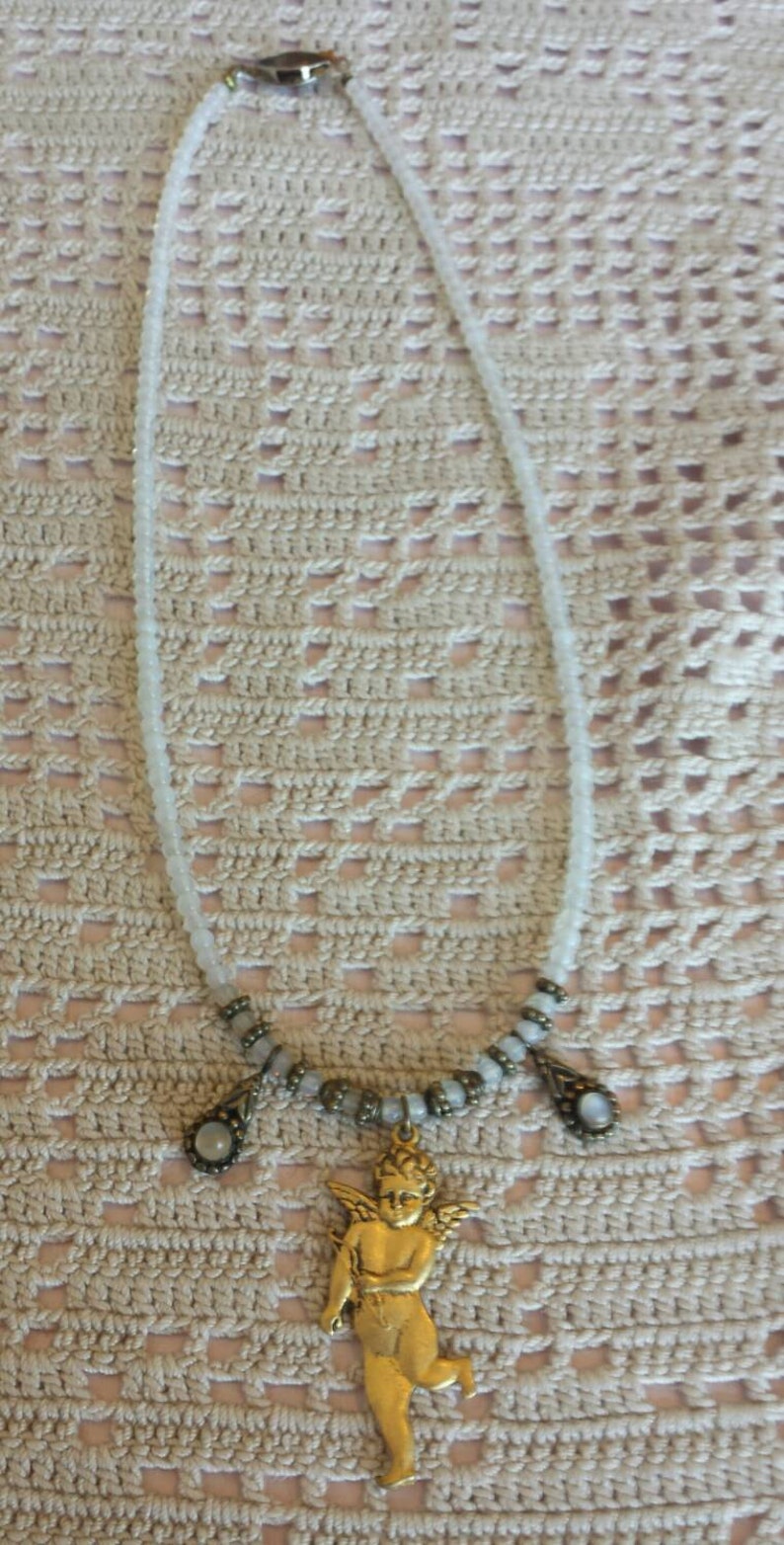 May include: A white beaded necklace with a gold cherub pendant and two smaller gold accents with white stones. The necklace is on a crocheted surface.