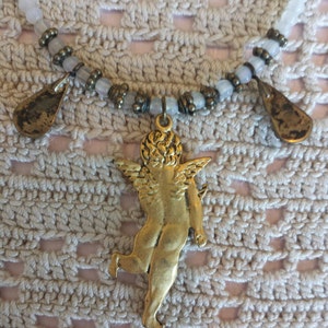 May include: A gold-toned necklace with a cherub pendant and two teardrop-shaped charms. The necklace is made with white and brown beads.