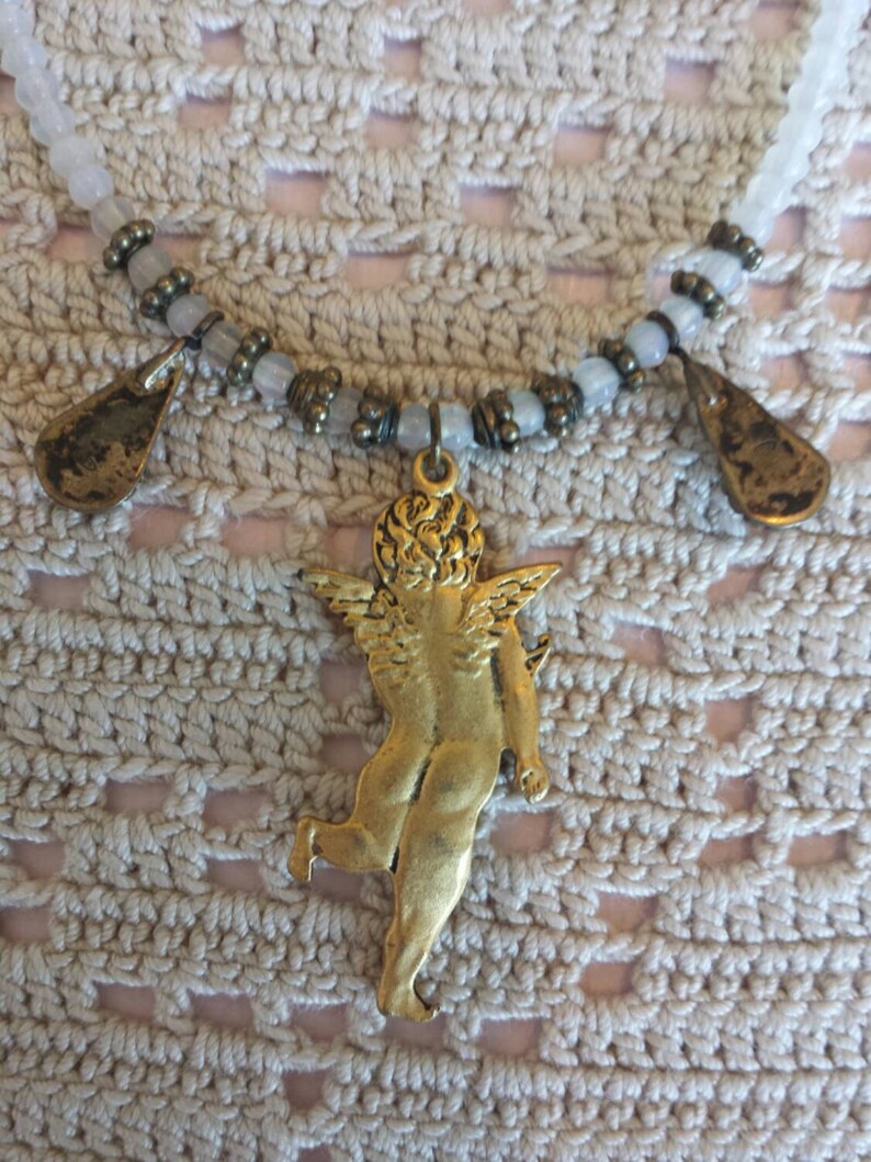May include: A gold-toned necklace with a cherub pendant and two teardrop-shaped charms. The necklace is made with white and brown beads.