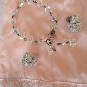 May include: A set of iridescent crystal jewelry featuring a bracelet, a pair of earrings, and a brooch. The jewelry is displayed on a pink satin fabric with white lace trim.