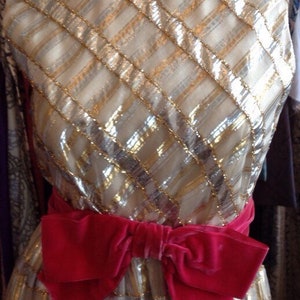 K&ouml;nnte beinhalten: Ein gold- und silberfarbenes Paillettenkleid mit einer gro&szlig;en rosa Schleife aus Samt an der Taille.