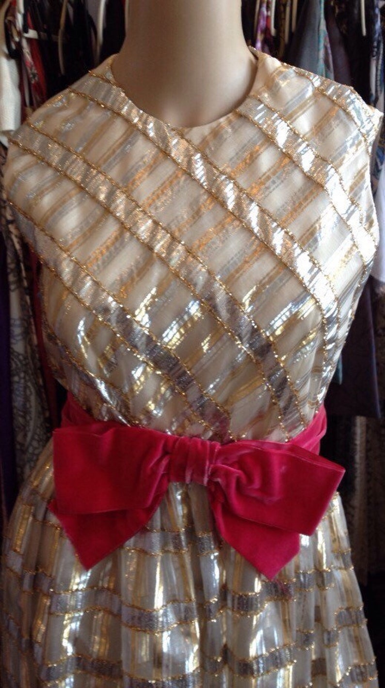K&ouml;nnte beinhalten: Ein gold- und silberfarbenes Paillettenkleid mit einer gro&szlig;en rosa Schleife aus Samt an der Taille.