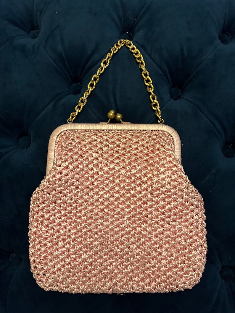 May include: A pink crocheted clutch bag with a gold chain strap. The bag has a clasp closure and a textured woven design.