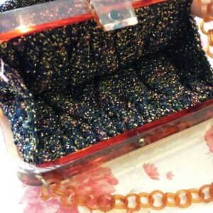 May include: A vintage handbag with a dark blue, glittery knit exterior. The frame is a translucent reddish-brown, with a matching chain strap. The interior is visible, showing the bag's capacity. The clasp is a clear, rectangular shape.
