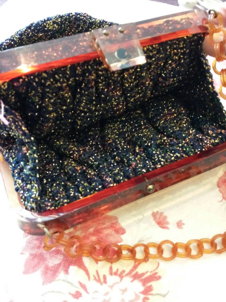 May include: A vintage handbag with a dark blue, glittery knit exterior. The frame is a translucent reddish-brown, with a matching chain strap. The interior is visible, showing the bag's capacity. The clasp is a clear, rectangular shape.