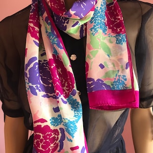 Puede incluir: Un pañuelo de seda blanco con estampado floral en tonos rosa, morado, azul y verde. El pañuelo tiene un borde rosa y la palabra "ECHO" está impresa en la parte inferior.