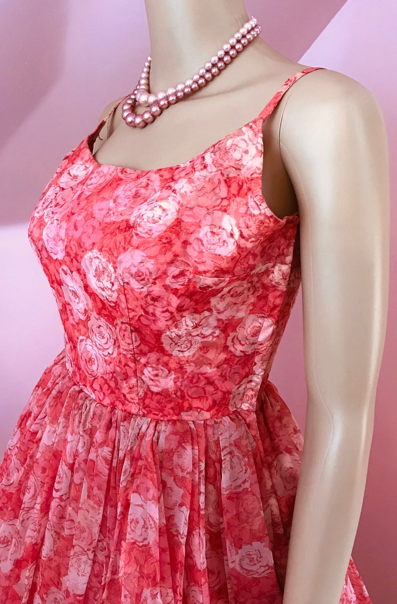 K&ouml;nnte beinhalten: Ein vintage-Kleid in rosa und wei&szlig; mit Blumenmuster, runder Ausschnitt und d&uuml;nnen Tr&auml;gern. Das Kleid ist aus einem durchsichtigen Stoff und hat einen vollen Rock.