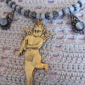 May include: A gold cherub pendant with wings, holding an arrow, hangs from a white beaded necklace with two teardrop-shaped charms.