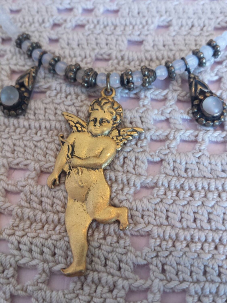May include: A gold cherub pendant with wings, holding an arrow, hangs from a white beaded necklace with two teardrop-shaped charms.