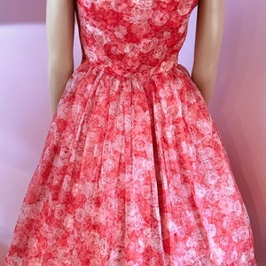 K&ouml;nnte beinhalten: Ein korallenfarbenes Kleid mit wei&szlig;em Blumenmuster. Das Kleid hat ein tailliertes Oberteil und einen vollen Rock. Es ist &auml;rmellos und hat einen runden Ausschnitt.