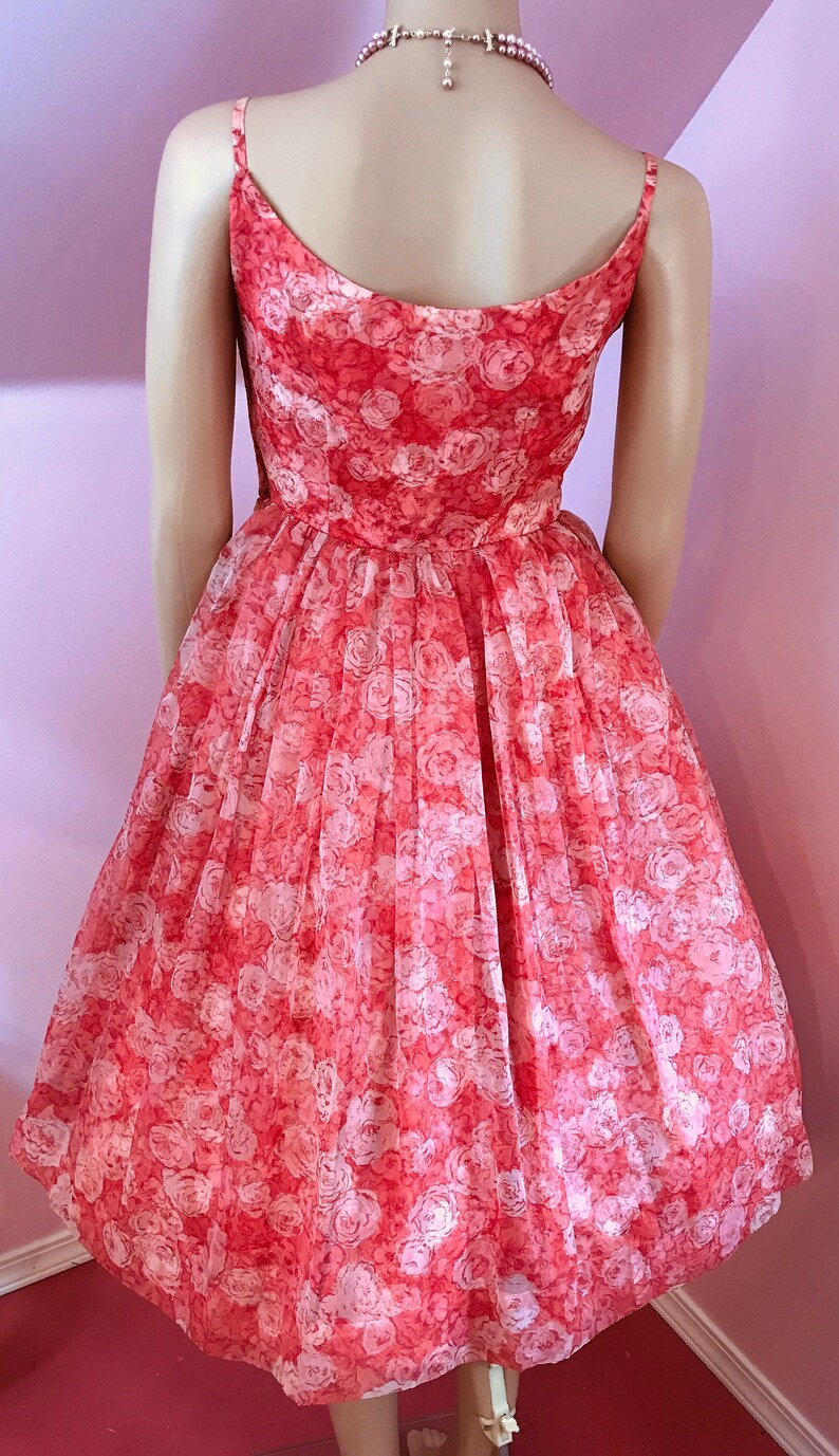 K&ouml;nnte beinhalten: Ein korallenfarbenes Kleid mit wei&szlig;em Blumenmuster. Das Kleid hat ein tailliertes Oberteil und einen vollen Rock. Es ist &auml;rmellos und hat einen runden Ausschnitt.