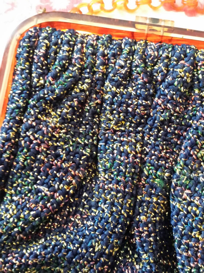May include: Close-up of a vintage navy blue clutch purse with a textured, woven design. The fabric is interwoven with threads of gold, pink, and green, creating a shimmering effect. The purse has a clear, rectangular frame.