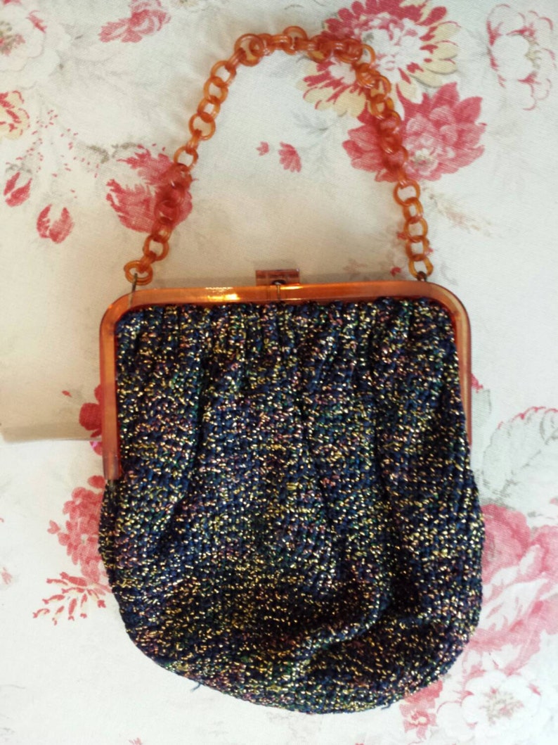 May include: Vintage evening bag with a shimmering, textured fabric in shades of blue, gold, and purple. The bag has a rectangular frame and a chain handle made of amber-colored plastic links. The clasp is a simple metal closure.