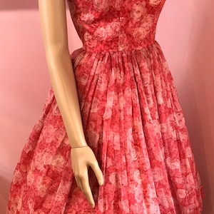K&ouml;nnte beinhalten: Ein pinkfarbenes, &auml;rmelloses Kleid mit einem Blumendruck aus wei&szlig;en Rosen. Das Kleid hat ein tailliertes Oberteil und einen vollen Rock.