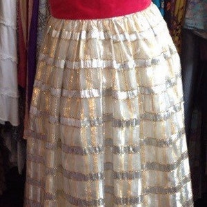 K&ouml;nnte beinhalten: Ein wei&szlig;es Kleid mit goldenen und silbernen Streifen und einer roten Samt-Sch&auml;rpe. Das Kleid ist aus einem gl&auml;nzenden Stoff gefertigt und hat ein tailliertes Oberteil und einen weiten Rock.