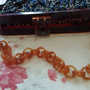 May include: A vintage brown acrylic handbag with a chain strap. The rectangular purse has a dark, textured top and a clear clasp. The chain is made of large, amber-colored links. The bag sits on a floral patterned surface.