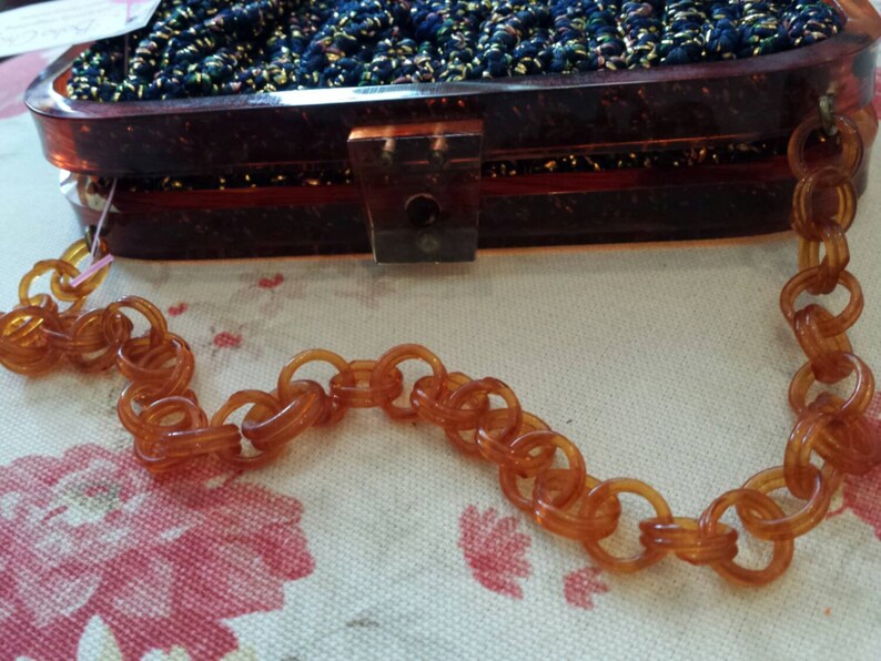 May include: A vintage brown acrylic handbag with a chain strap. The rectangular purse has a dark, textured top and a clear clasp. The chain is made of large, amber-colored links. The bag sits on a floral patterned surface.