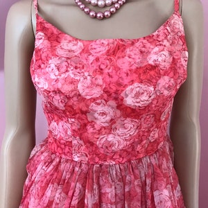 K&ouml;nnte beinhalten: Ein pinkfarbenes, &auml;rmelloses Kleid mit einem Blumendruck aus wei&szlig;en Rosen. Das Kleid hat ein tailliertes Oberteil und einen weiten Rock.